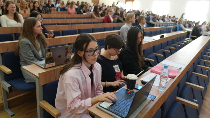 Счастливая молодая семья получает ключи от новой квартиры в студенческом общежитии, рядом стоят коробки с вещами и учебниками. На заднем плане видны окна здания.