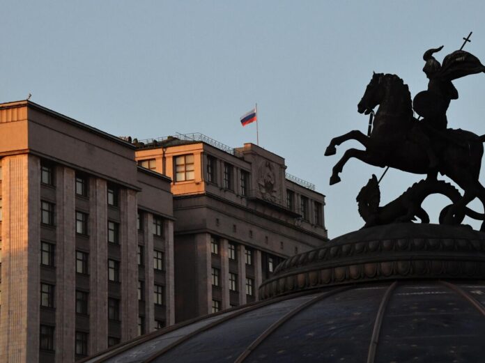 Искусственный интеллект в образе робота сидит за столом, как на допросе, напротив человека в деловом костюме, символизирующего Госдуму. На столе документы и микрофон.