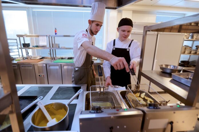 Студенты за работой на кухне, готовящие еду в необычном месте, напоминающем хоккейную коробку.